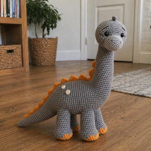 Puede incluir: Un juguete de dinosaurio tejido a mano en gris con detalles en naranja. El dinosaurio tiene un cuello largo, una cola y cuatro patas. La espalda presenta púas naranjas y los pies son naranjas. El juguete está sobre un suelo de madera.