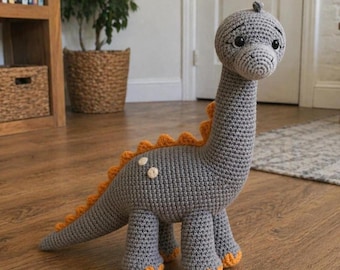 May include: A handmade crochet dinosaur toy in gray with orange accents. The dinosaur has a long neck, a tail, and four legs. The dinosaur's back has orange spikes, and its feet are orange. The toy is standing on a wooden floor.