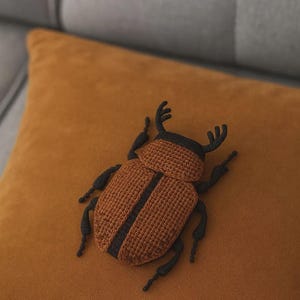 Könnte beinhalten: Ein handgefertigter, gehäkelter Käfer in Braun- und Schwarztönen, der auf einem senffarbenen Kissen liegt. Der Käfer hat ein detailliertes Design mit segmentiertem Körper und Beinen und ist wahrscheinlich ein Dekorationsartikel.