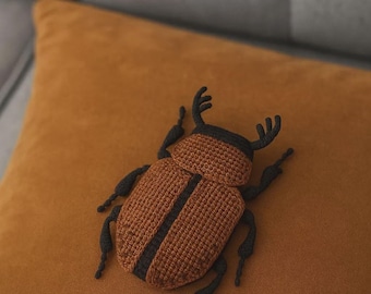 May include: A handmade, crocheted beetle in shades of brown and black, resting on a mustard-colored pillow. The beetle features a detailed design with segmented body and legs, and is likely a decorative item.