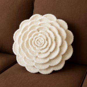 Puede incluir: Un cojín de ganchillo de color crema con forma de flor grande. El cojín tiene múltiples capas de pétalos, creando un efecto tridimensional. El cojín está colocado sobre un sofá marrón.