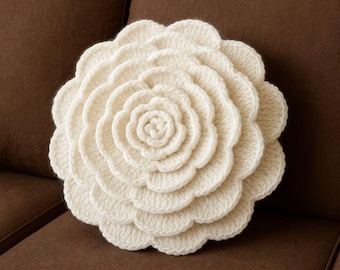 May include: A cream-colored, crocheted pillow shaped like a large flower. The pillow has multiple layers of petals, creating a three-dimensional effect. The pillow is placed on a brown sofa.