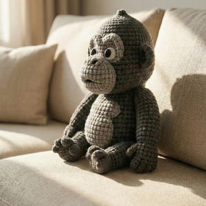 Puede incluir: Una mu&ntilde;eca de mono de ganchillo gris hecha a mano con cara, orejas y pecho de color gris claro. El peluche tiene ojos grandes y redondos y est&aacute; sentado en un sof&aacute; beige. El mono es un juguete suave y tierno.