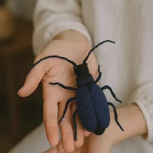 May include: A handmade, dark blue crochet beetle with black accents. The insect has six legs and long antennae. The beetle is held in an open hand against a soft, neutral background.