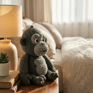 Puede incluir: Un mu&ntilde;eco de mono de ganchillo gris hecho a mano, con la cara y el vientre de color gris claro, est&aacute; sentado en una mesita de noche de madera. El mono tiene ojos grandes y est&aacute; junto a una l&aacute;mpara, libros y una peque&ntilde;a planta en una maceta blanca.