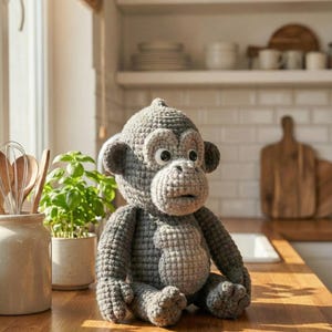 Puede incluir: Una mu&ntilde;eca de mono de ganchillo gris hecha a mano con ojos grandes y una expresi&oacute;n de sorpresa. El peluche est&aacute; sentado sobre una superficie de madera, con un fondo de cocina con plantas y estantes.