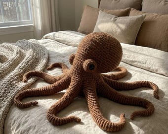 May include: A large, brown crocheted octopus plush toy with eight long, curved tentacles. The octopus has a round head with two small, dark eyes. It is lying on a bed with neutral-colored bedding and pillows. A beige crocheted blanket is visible.