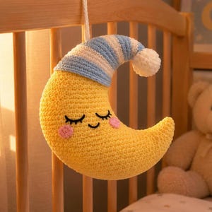 May include: A yellow crescent moon plush toy with a crocheted texture, featuring a sleeping face with closed eyes and pink cheeks. The moon wears a blue and white striped nightcap with a white pom-pom at the tip, suspended by a white cord.