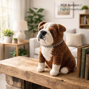 Puede incluir: Un perro bulldog tejido a ganchillo, marrón y blanco, con un collar de cadena plateada, sentado sobre una mesa de madera. El texto "PDF Pattern / Digital Download" es visible en el fondo.