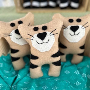 May include: Three tan and black stuffed tiger toys with white faces and black whiskers sit on a teal patterned fabric. Additional tiger toys are visible in the background, inside a wooden crate. The toys have black stripes.