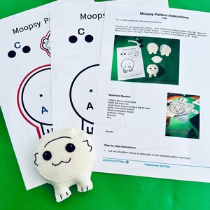 May include: A felt Moopsy character with black eyes and a stitched mouth sits on a green surface next to pattern instructions. The instructions include a list of materials and step-by-step instructions. The text "Moopsy Pattern" is visible.