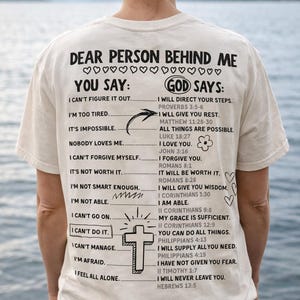 May include: Cream-colored t-shirt with black text. The shirt features the phrases "Dear Person Behind Me, You Say" and "God Says" with a list of statements and corresponding Bible verses. A cross and hearts are also included.