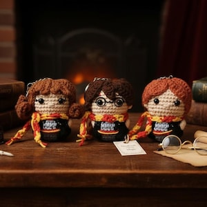 Op de afbeelding: Drie handgemaakte gehaakte poppen met bruin, rood en rossig haar, elk met een gele en rode sjaal. De poppen hebben zwarte outfits en worden tentoongesteld op een houten oppervlak met boeken, een veer en een bril.