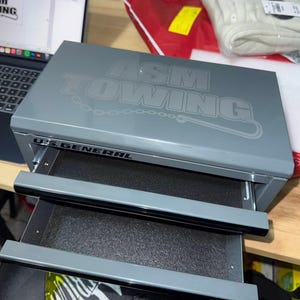 May include: A gray metal tool chest with two open drawers. The top of the chest has the text "ASM TOWING" in a lighter gray font. The brand "U.S. GENERAL" is printed on the front of the chest.