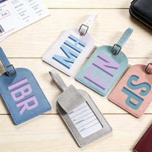 May include: A collection of personalized luggage tags in various pastel colors, including blue, pink, and gray. Each tag features initials in a contrasting color and includes a strap with a buckle. A boarding pass and wallet are in the background.