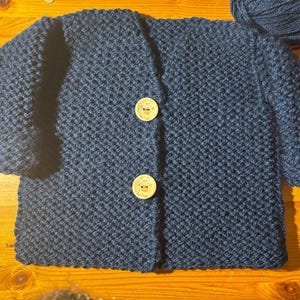 May include: A navy blue, hand-knitted cardigan with two large, light-colored buttons. The textured knitwear features a V-neck design and long sleeves. A ball of matching yarn is visible in the background. This cozy, handmade garment is perfect for babies and toddlers.