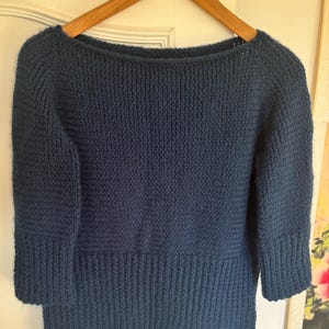 May include: A dark blue knitted sweater with a wide boat neckline and three-quarter length sleeves. The garment features a textured knit pattern on the main body and a vertical ribbed knit design on the cuffs and the lower hemline of the sweater.
