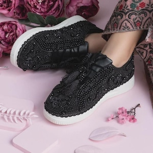 Pode incluir: Tênis pretos com contas e solas brancas. Os tênis apresentam intrincados trabalhos de contas e borlas decorativas. Os sapatos estão em uma superfície rosa com detalhes florais.