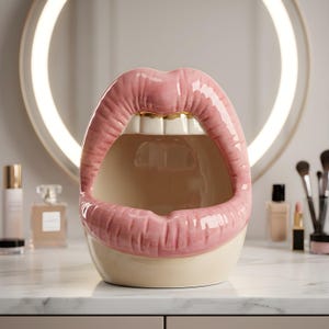May include: A ceramic container shaped like an open mouth with glossy pink lips and white teeth. The interior is hollow and cream-colored, featuring a gold-toned accent on the upper teeth. The object has a smooth, reflective finish throughout.