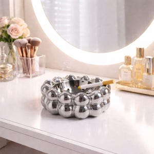 May include: A silver ashtray with a unique bubble design. The ashtray holds a lit cigarette. The ashtray is on a white surface, with a mirror and beauty products in the background. The ashtray is a decorative item.