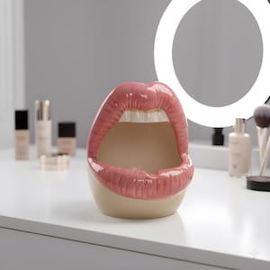 Puede incluir: Un recipiente de cerámica con forma de labios con un diseño de labios rosa brillante y dientes blancos. El interior es de color beige neutro. El recipiente está sobre una superficie blanca, con productos de maquillaje y una luz de anillo en el fondo.