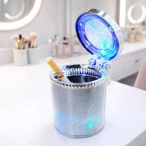 May include: A silver and clear ashtray with a cigarette inside. The ashtray has a decorative silver trim and a blue light. The lid is open, revealing a blue light design. The ashtray is on a white surface.
