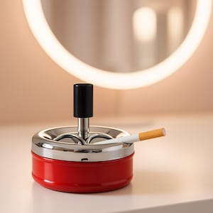 May include: A red and chrome ashtray with a black handle and a cigarette resting on the edge. The ashtray is round and sits on a white surface. A lit cigarette is in the ashtray.