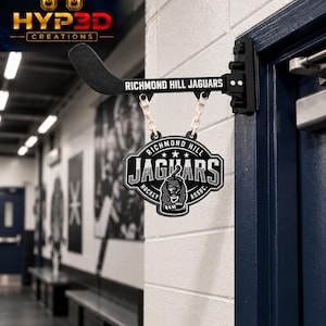 May include: A black wall-mounted hockey stick bracket with the text Richmond Hill Jaguars, featuring a hanging sign with the Richmond Hill Jaguars Hockey Assoc. logo, a jaguar graphic, and white plastic chain links attached to the bracket.