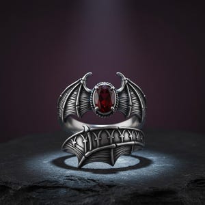 Könnte beinhalten: Ein silberner Ring mit einem Fledermausflügel-Design und einem roten, ovalen Edelstein. Die Flügel des Rings sind mit geätzten Linien detailliert, und der Edelstein ist in einer dekorativen Lünette gefasst. Der Ring wird auf einer dunklen Oberfläche präsentiert.