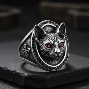 Puede incluir: Anillo plateado con un detallado rostro de gato con ojos de piedras preciosas rojas. El gato está engastado en un marco ovalado, rodeado de piedras negras. Un diseño de cruz decorativa adorna la banda del anillo.