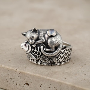 Puede incluir: Anillo de plata con un gato durmiendo sobre un pez. El anillo tiene un diseño detallado y texturizado, con una pequeña piedra redonda de color claro incrustada en la espalda del gato. La banda tiene un patrón de olas.