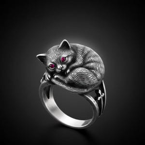 Puede incluir: Anillo de plata con un diseño detallado de un gato enroscado con ojos de piedras preciosas rojas. El anillo tiene un acabado texturizado y una banda decorativa. El pelaje del gato está intrincadamente tallado, sobre fondo oscuro.