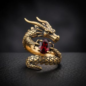 May include: A gold-colored dragon ring with a red gemstone held in its claws. The dragon's body wraps around the finger, with detailed scales and a textured finish. The ring is set against a dark background.