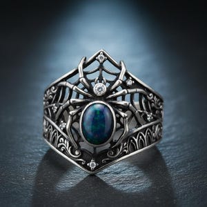 May include: A silver ring featuring a spider design with an oval, dark blue gemstone at its center. The ring has a web-like pattern and is adorned with small, clear gemstones. The ring's design is intricate and detailed.