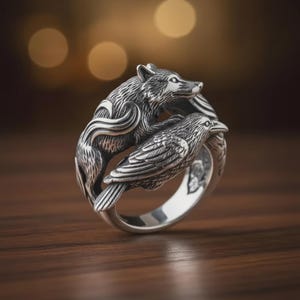 May include: A silver ring featuring a detailed wolf and raven design. The wolf is positioned above the raven, with intricate carvings on both animals. The ring has a polished finish and is set against a blurred background.