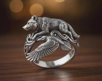 May include: A silver ring featuring a wolf leaping over a raven with outstretched wings. The ring has intricate detailing, with the wolf and raven sculpted in a realistic style. The ring is set against a blurred background.