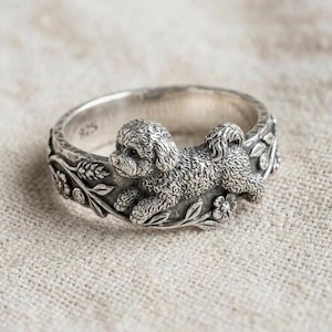 May include: A silver ring featuring a detailed sculpture of a dog lying down. The ring band is adorned with floral and leaf patterns, with small, sparkling accents. The dog's fur is textured, and the ring has a polished finish.