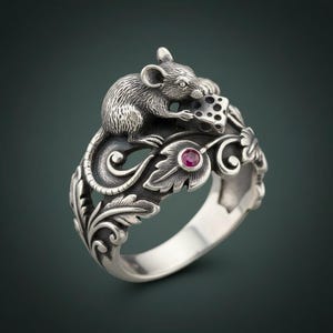 Puede incluir: Anillo de plata con un ratón detallado sosteniendo un trozo de queso. El anillo tiene un diseño floral ornamentado y una pequeña gema roja redonda. El anillo está sobre un fondo oscuro.