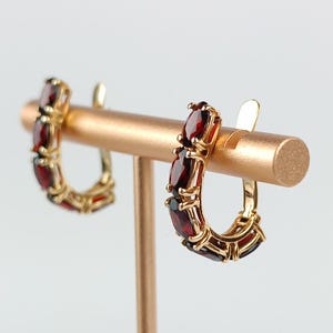 May include: A pair of gold hoop earrings featuring a series of deep red, oval-cut garnet gemstones set in a prong setting. The earrings are curved in a semi-circular shape, showcasing the polished gold metal frame and the faceted dark red stones.