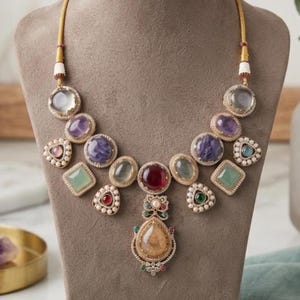 Könnte beinhalten: Eine goldfarbene Halskette mit einem zentralen tropfenförmigen Steinanhänger, umgeben von verschiedenen bunten Cabochon-Steinen in runden, quadratischen und dreieckigen Formen. Die Steine sind mit kleinen weißen Perlen und klaren Kristallen verziert.
