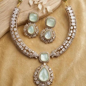 May include: A jewelry set consisting of a necklace and matching earrings. The necklace features a large, central pear-shaped light green stone surrounded by clear crystals, with a curved band of clear crystals. Two matching pear-shaped earrings.
