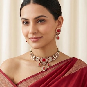May include: A gold-toned choker necklace and matching drop earrings set featuring deep red rectangular and teardrop-shaped stones. The jewelry is intricately detailed with small white accents and ornate metalwork, creating a cohesive set.