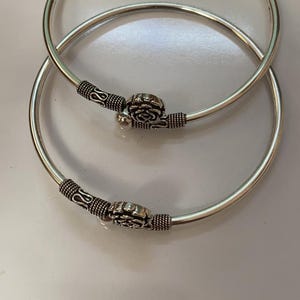 May include: Two silver-toned metal bangles featuring a smooth, circular band design. Each bangle is accented with a central floral motif, flanked by textured, coiled metal detailing on both sides of the flower, creating a consistent look.