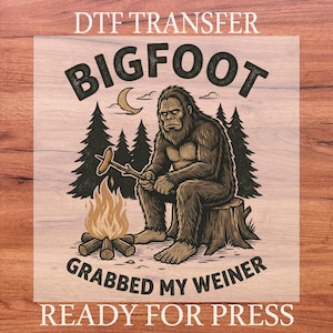 May include: DTF transfer featuring a Bigfoot illustration. The design shows Bigfoot sitting by a campfire, holding a sausage on a stick. Text includes "BIGFOOT GRABBED MY WEINER" and "READY FOR PRESS."