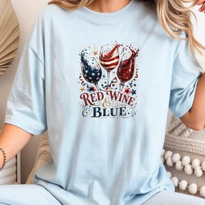 May include: A light blue t-shirt featuring a graphic of three wine glasses filled with red, white, and blue liquid, surrounded by stars and confetti. The text "RED WINE & BLUE" is printed in a decorative font below the wine glasses.