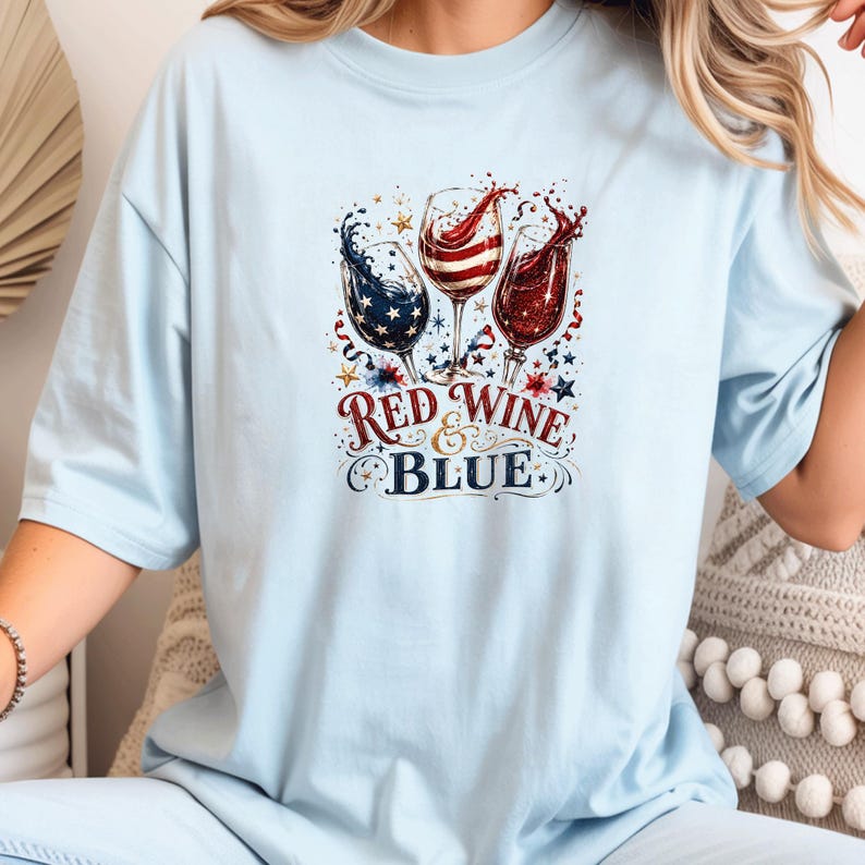 May include: A light blue t-shirt featuring a graphic of three wine glasses filled with red, white, and blue liquid, surrounded by stars and confetti. The text "RED WINE & BLUE" is printed in a decorative font below the wine glasses.