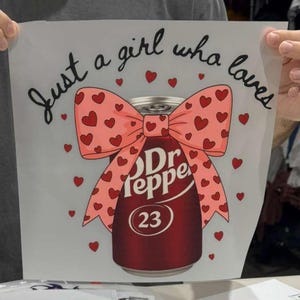 May include: A white sheet featuring a graphic of a maroon Dr Pepper soda can adorned with a large pink bow covered in small red hearts. The text "Just a girl who loves" is printed in black cursive above the can, surrounded by scattered red hearts.
