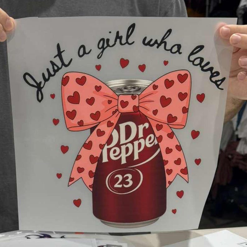 May include: A white sheet featuring a graphic of a maroon Dr Pepper soda can adorned with a large pink bow covered in small red hearts. The text "Just a girl who loves" is printed in black cursive above the can, surrounded by scattered red hearts.