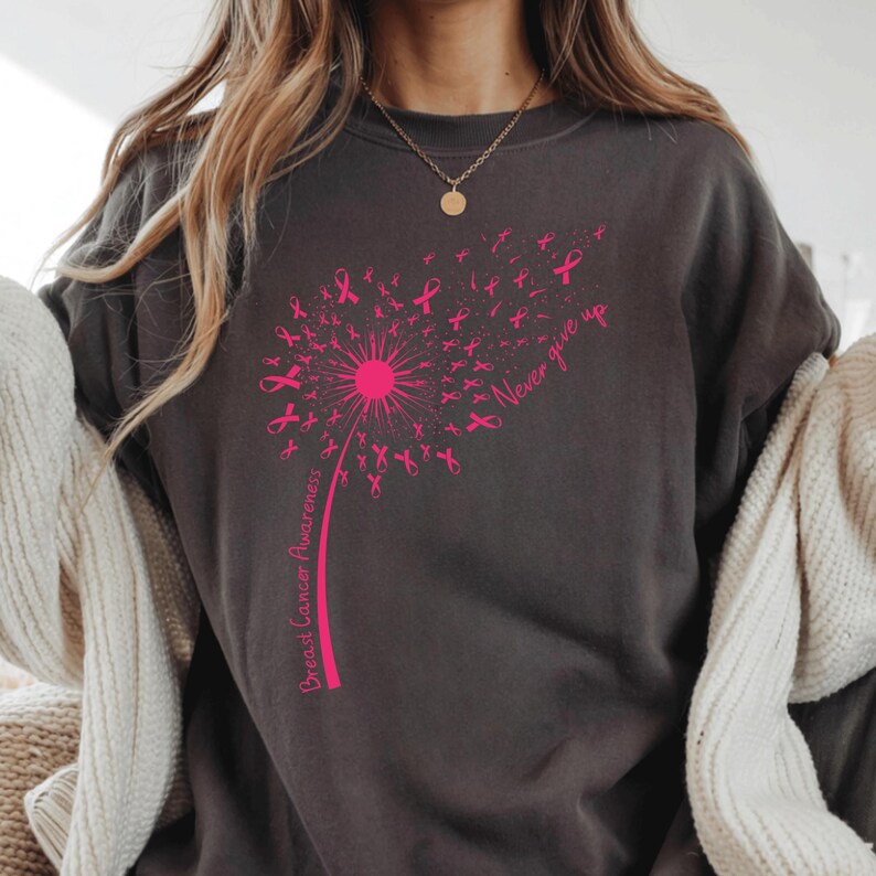 May include: A charcoal gray crewneck sweatshirt featuring a bright pink graphic of a dandelion where the seeds are shaped like breast cancer awareness ribbons. The text "Breast Cancer Awareness" and "Never give up" are printed in pink.