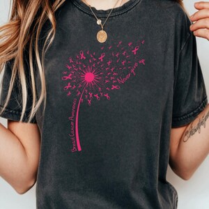 May include: A charcoal gray t-shirt featuring a pink graphic of a dandelion where the seeds are small breast cancer awareness ribbons. The text "Breast Cancer Awareness" runs along the stem, and "Never give up" is printed near the top right.